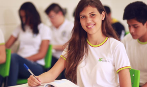 Aluna, com o uniforme do IFTM, na sala de aula. Ela está segurando uma caneta, ao seu redor há outros alunos.